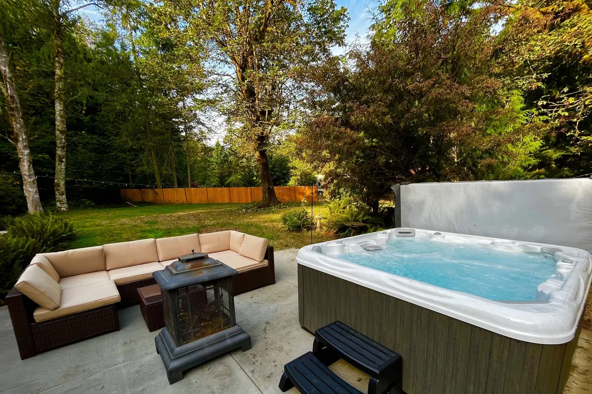 This image features a hot tub nestled among surrounding trees, an outdoor seating area with comfortable furniture, and a nearby fire pit. The space is enveloped in natural greenery, creating a tranquil and welcoming atmosphere perfect for relaxation and social gatherings.