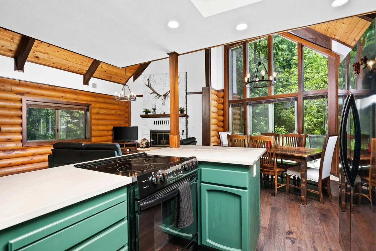 A beautiful kitchen and dining area with a central island, large windows looking out to greenery, exposed wooden beams, and a view toward a stone-accented fireplace. The space looks both modern and rustic, with ample seating—ideal for family meals and entertaining.