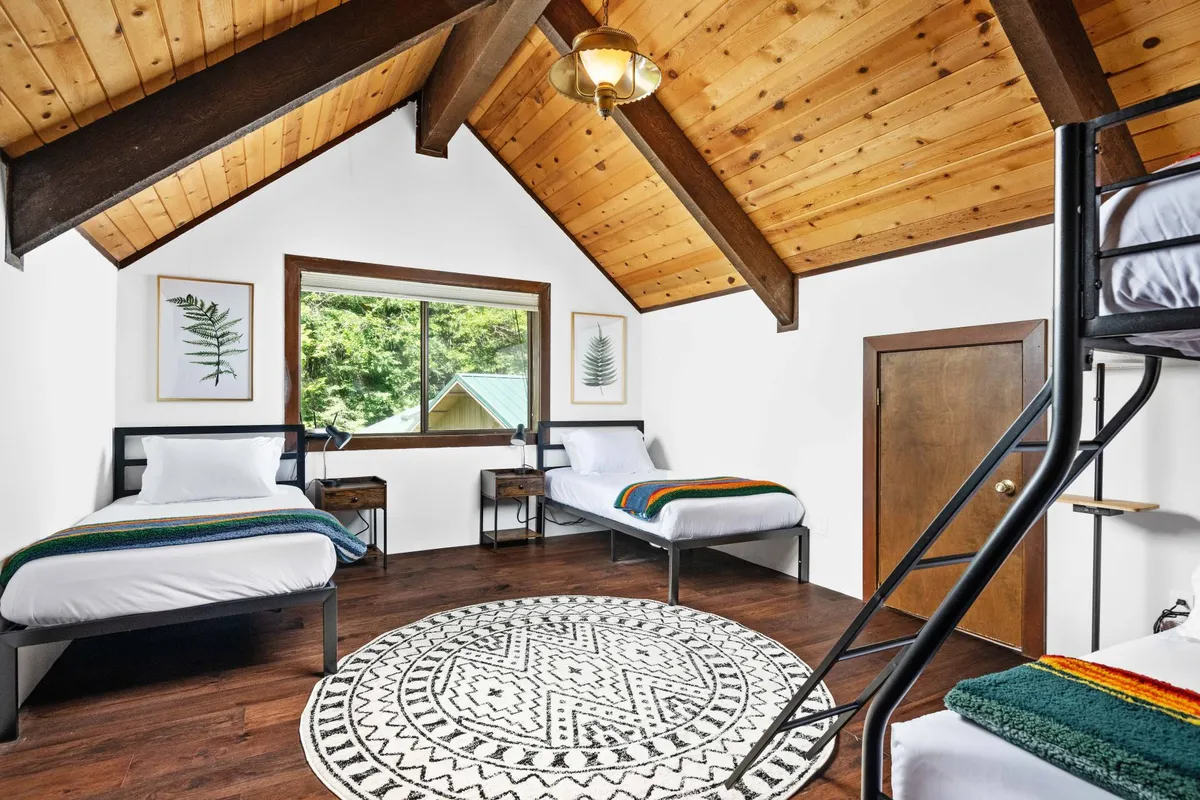 A cozy upstairs space highlighted by a wooden bunk bed, decorative rug, exposed wooden beams, and a window with views of greenery. The setup is playful and communal, inviting for children or young adults.