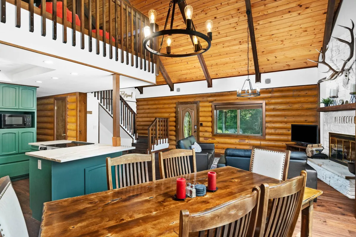 A wide-angle interior view that highlights the open-plan design connecting the dining table, kitchen island, and living area. Wooden beams and a fireplace enhance the rustic charm, while large windows draw the outdoors in. The overall arrangement underscores spaces for cooking, dining, and relaxing together.