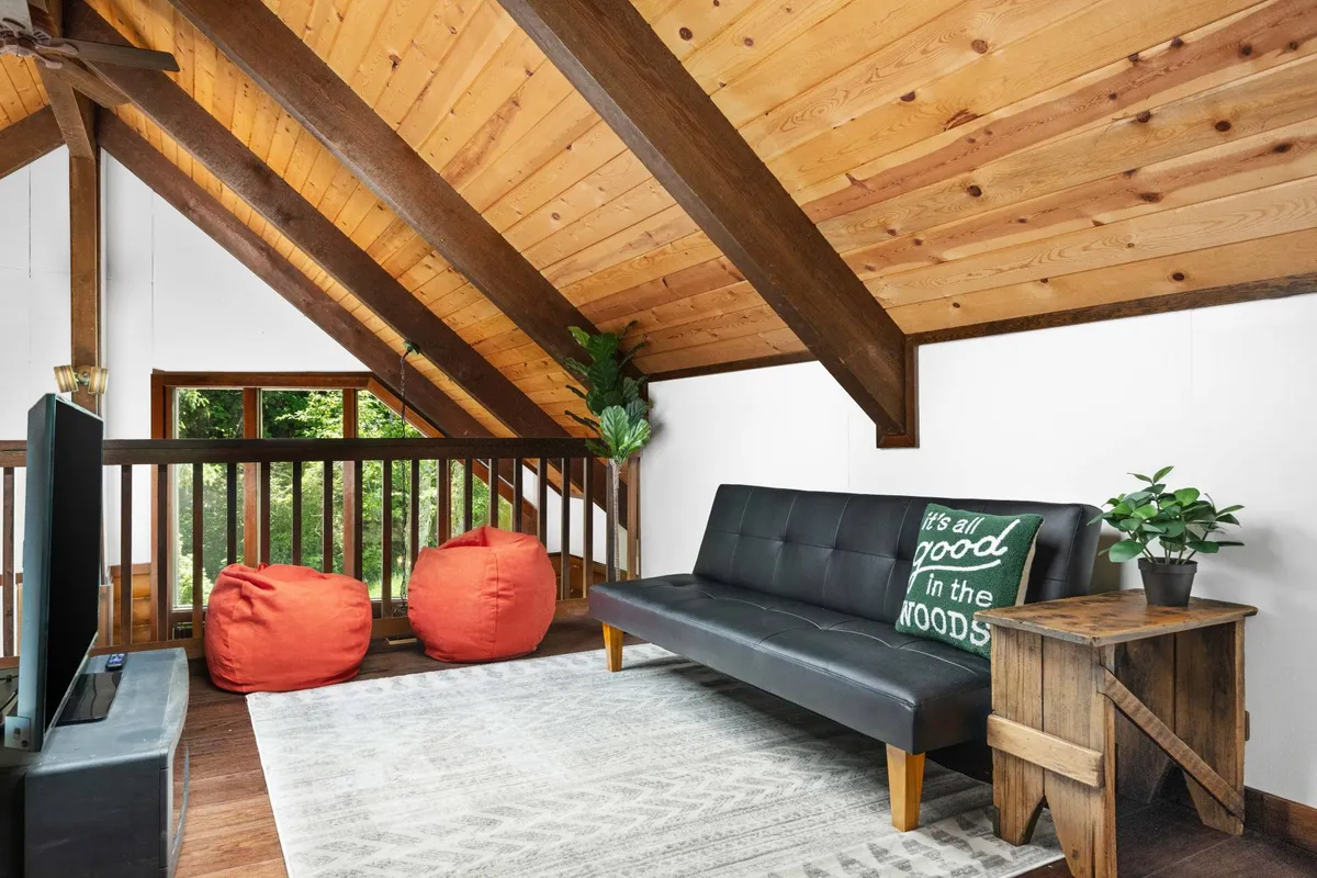 A comfortable living/sleeping combo featuring a sleek black sofa, two vibrant orange bean bags, rustic wooden beams, and a TV. The room is versatile, designed for both lounging and sleeping, with fun touches ideal for group activities or extra guests.