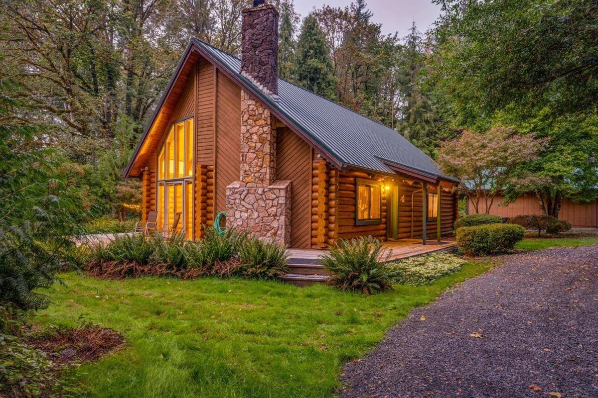 This image captures the log cabin exterior with a prominent stone chimney, expansive windows, and a landscaped garden, all set against a wooded backdrop. The setting evokes the spacious, natural grounds that make The Fernwood ideal for pets to explore and owners to enjoy outdoor relaxation.