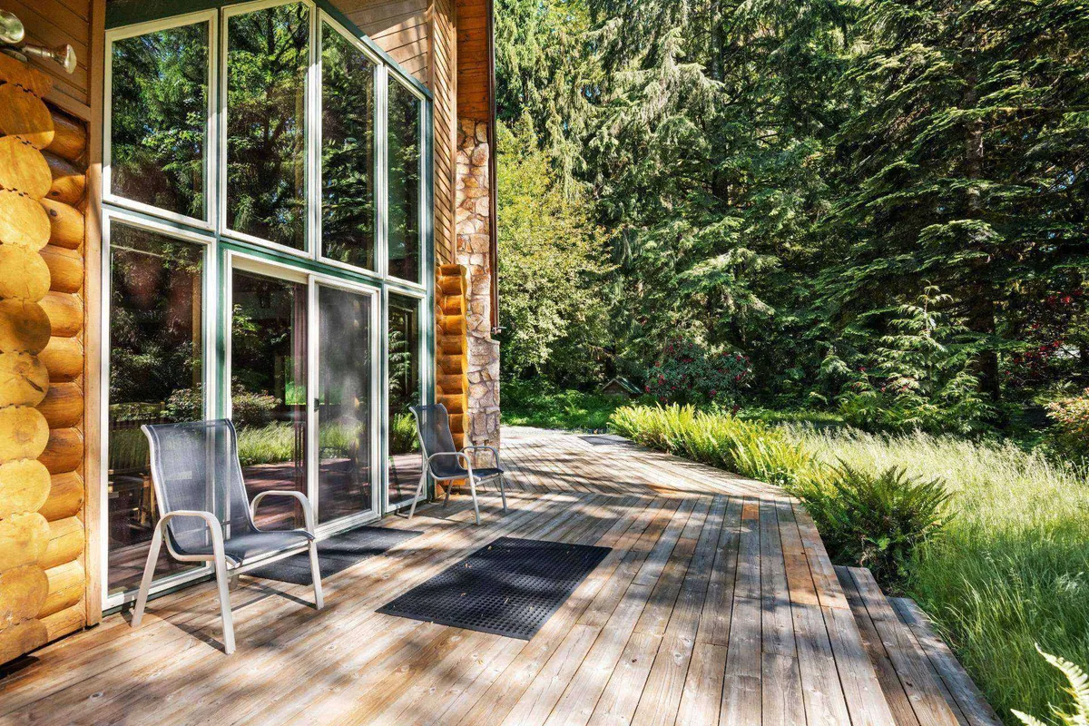 A wide wooden deck extends from the house, framed by large glass windows and embraced by dense greenery. The setting is ideal for enjoying coffee outdoors, socializing, or simply soaking in the sights and sounds of nature.