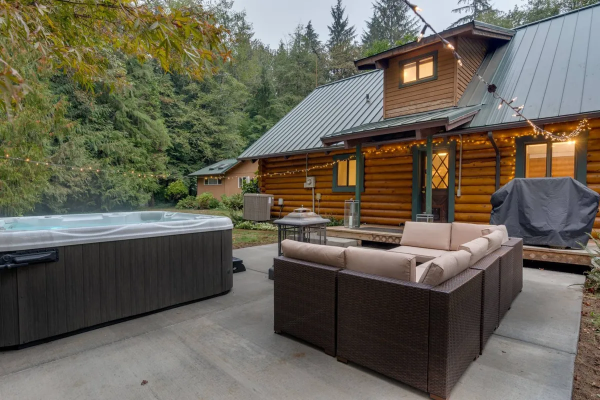 The log cabin's exterior with a spacious outdoor area featuring a large hot tub and ample seating, set amidst trees. The space is inviting for relaxation and family gatherings after a day of adventure.