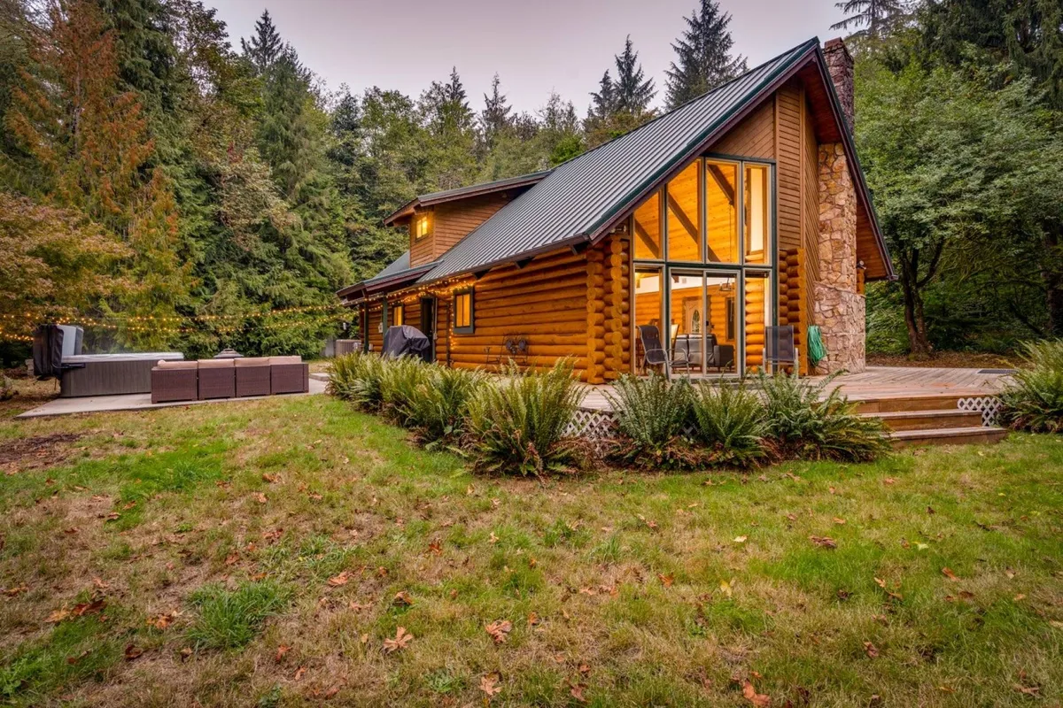 The image showcases the exterior of a modern log cabin nestled among lush greenery, featuring a charming deck with outdoor seating and expansive windows that invite sunlight into the home. The tranquil setting hints at opportunities for outdoor dining, relaxing amidst nature, and gathering around a fire pit as evening falls.