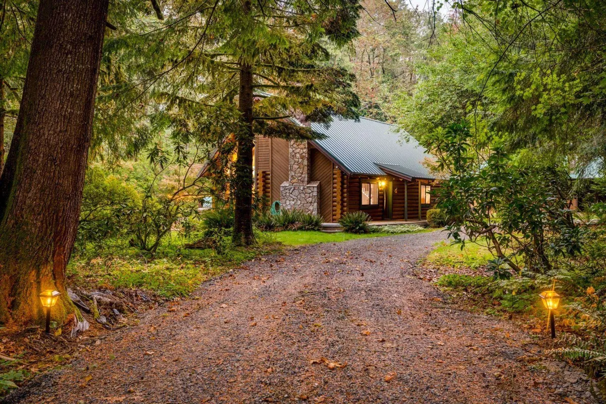 A log cabin with a striking stonework facade sits surrounded by nature, with a lantern-lit pathway leading to the entrance. The setting exudes a tranquil, welcoming vibe that hints at both relaxation and adventure. The surrounding trees and natural landscaping suggest proximity to outdoor activities.