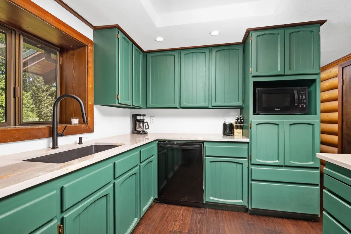 A cozy, renovated kitchen featuring green cabinetry, black appliances, a marble countertop, and a wooden window frame. The space feels inviting and practical, perfect for preparing meals or gathering for coffee. Sunlight streams in, highlighting the thoughtful updates and homey touches.