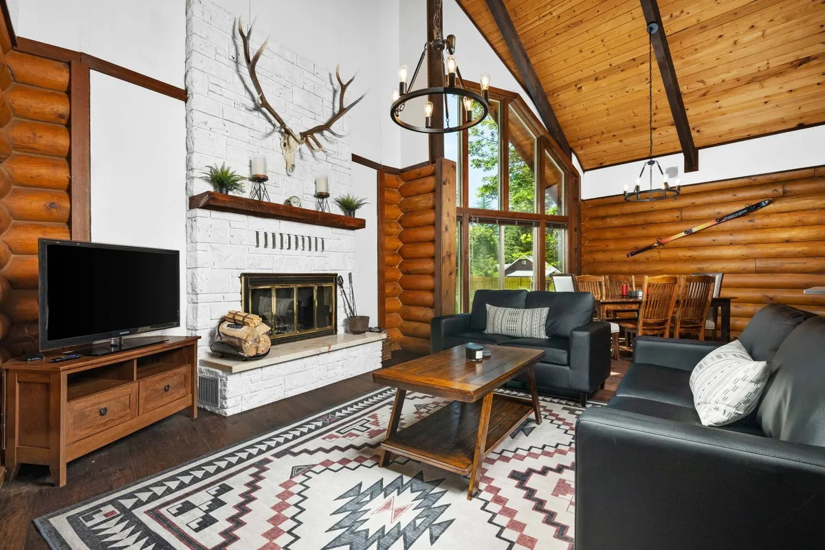 This image captures a spacious, open-concept living area featuring a prominent stone fireplace, large windows that flood the room with natural light, and a cozy arrangement of sofas and a rug. The adjacent dining area is visible, hinting at the seamless flow between living and dining spaces. Rustic and modern décor elements blend to create a warm, inviting atmosphere perfect for gatherings.