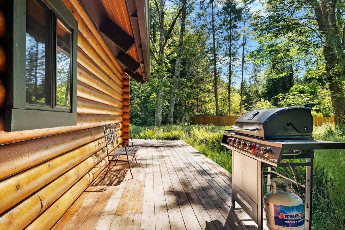 This image showcases the log cabin exterior, a grill, a spacious wooden deck, and lush surrounding greenery. The scene radiates tranquility, with the deck appearing as an ideal spot for evening relaxation, outdoor dining, or stargazing after a day of adventure.
